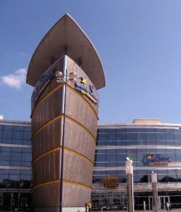 Bronzener Turm in Dreiecksform vor einem riesigen Gebäude mit großer Glasfront.