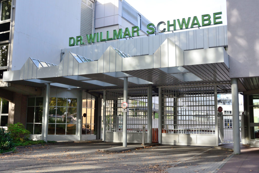 Eingangsbereich eines großen Gebäudes mit zwei großen quadratischen, geschlossenen Metalltoren. In großen grünen Buchstaben steht darüber: "WILLMAR SCHWABE"