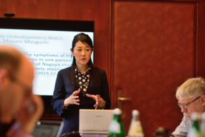 Frau stehend vor einem Tisch mit Laptop. Im Hintergrund eine Leinwand mit englischem Text. Am Tisch nur angeschnitten zu erkennen, weitere Menschen sitzend.