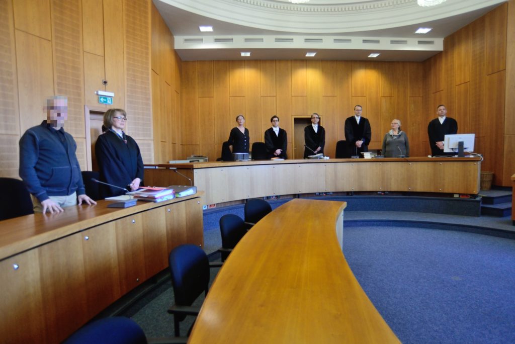 Gerichtssaal von innen, hölzerne Wandvertäfelung, blauer Teppich. An zwei langen Pulten stehen Richter:innen und Anwält:innen.