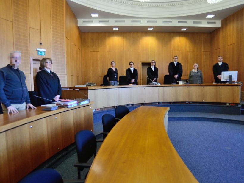 Gerichtssaal von innen, hölzerne Wandvertäfelung, blauer Teppich. An zwei langen Pulten stehen Richter:innen und Anwält:innen.