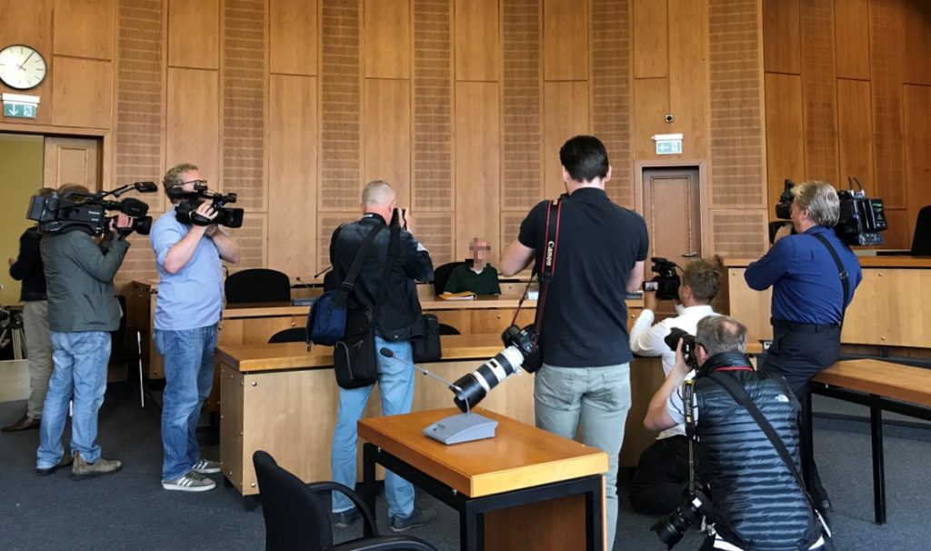 Gerichtssaal mit holzvertäfelten Wänden, viele Reporter mit Kameras auf die Anklagebank gerichtet.