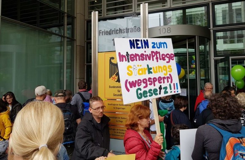 Gruppe protestierender Menschen vor einem Gebäude-Eingang. Eine Frau hält Schild in der Hand mit der Aufschrift: "NEIN zum IntensivPflegeStärkungs(wegsperr)Gesetz!!!!"