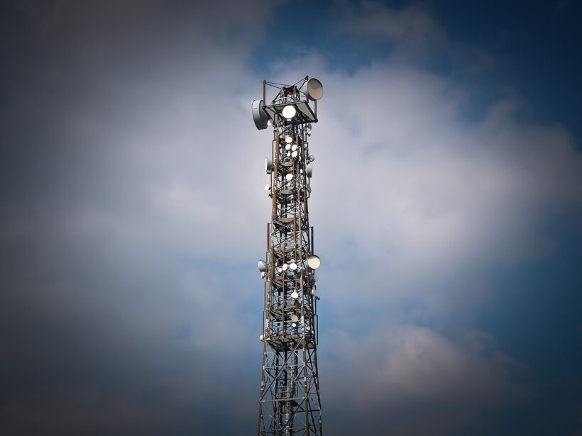 Funkturm aus Stahl, mit Antennen und Sat-Schüsseln; vor einem dunkelblauen Himmel mit weißen Wolken.