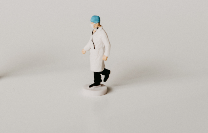 Foto: Ein Arzt mit blauer Haube und Mundschutz, nicht genau zu erkennen, steht auf einer weißen Fläche.