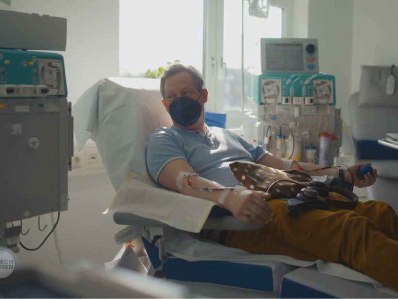 Mann mit FFP2-Maske vor Nase und Mund in einem Krankenhauszimmer auf einer Liege. Sein Arm ist voller Zugänge mit Schläuchen. Medizinische Geräte um ihn herum.