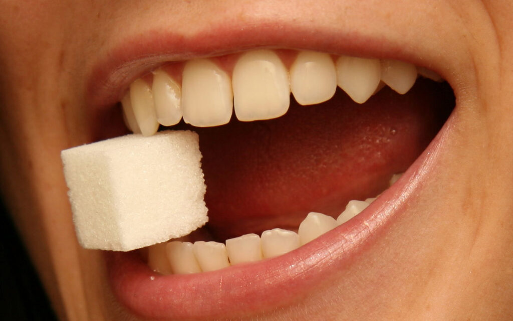 geöffneter weiblicher Mund; die Zähne halten einen Zuckerwürfel.